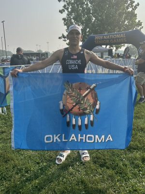 Man standing in front of National Senior Games Triathlon finish line smiling and holding Oklahoma flag.