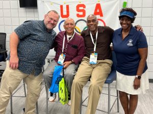 Four people smile and pose together in front of a USA Olympic flag.