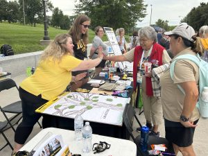 An outdoor fair setting with tables displaying information. A woman on one side of a table high fives a woman on the other side.