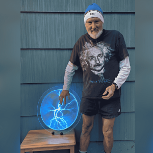 Glenn Govertsen stands with his hand on a plasma disc with radiating lines used during his physics demonstrations. Glenn is dressing in a tshirt with Albert Einstein on it, athletic shorts, and a winter cap.
