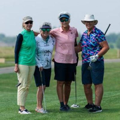 Four golfers holding golf clubs stand together on the green smiling.