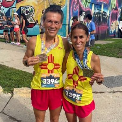 Tim and Rigil Vigil smile together while each holding a medal. They are wearing matching red shots and yellow tank tops with the New Mexico flag.