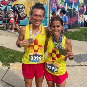 Tim and Rigil Vigil smile together while each holding a medal. They are wearing matching red shots and yellow tank tops with the New Mexico flag.