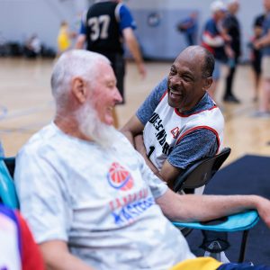 Two older men in athletic wear sit across from each other next to a basketball court and share a laugh.