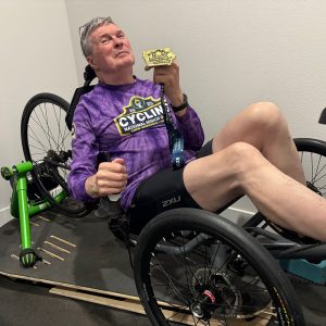 Jim DeGoey sits on his recumbent bike while holding the gold medal he won at the 2025 National Senior Games.