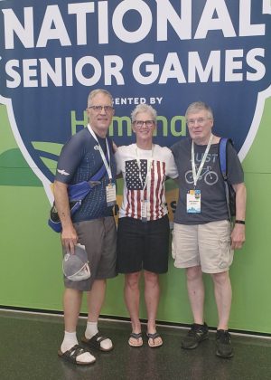 Three people stand in front of a large logo decal for the 2025 National Senior Games.