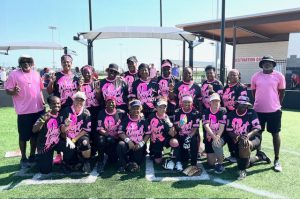 A group photo of a women's softball team at the National Senior Games with their two coaches. They are wearing pink and black uniforms and posed together on a softball field.