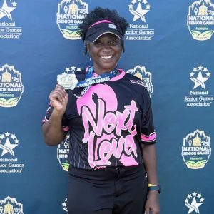 Brenda Jones, a 65-year-old woman, smiles while holding a medal earned at the 2025 National Senior Games presented by Humana. She is wearing a pink and black softball uniform with the words, "Next Level," on the front.