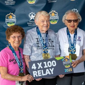 Three older women athletes stand together for an awards picture, holding a 4x100 Relay sign and wearing gold medals.