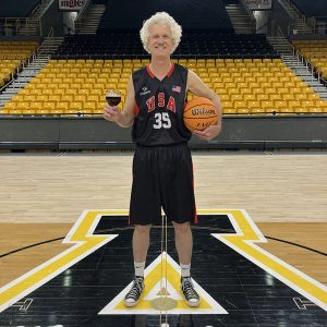 Kinney Baughman stands on a basketball court wearing a basketball uniform. He is holding a glass of beer in one hand and a basketball in the other.