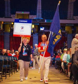 Bonita Robbins and another athlete lead the Kansas delegation at the National Senior Games Parade of Athletes holding a sign and the Kansas flag.
