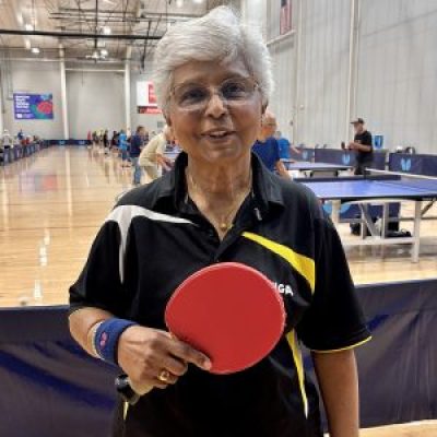 Sylvana Airan poses holding her table tennis paddle. Behind her is a long row of table tennis tables and people playing.
