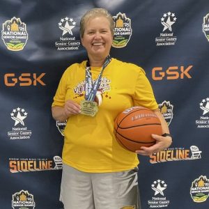 Kari Torkelson smiles in front of a background for the 2025 National Senior Games holding a basketball in one hand and a gold medal in the other.
