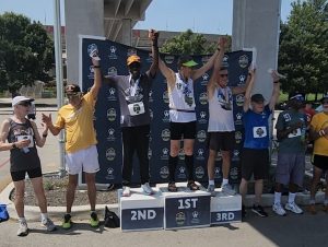 A group of winners at the National Senior Games posing on and around a podium with their arms lifted above their heads in celebration.