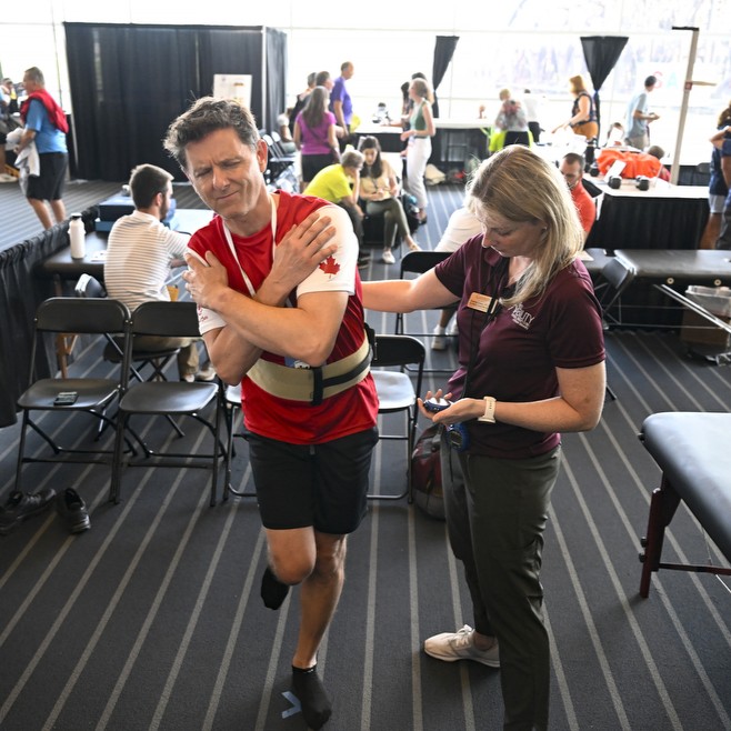 An athlete balances on one leg with eyes closed as part of the Sustained Athlete Fitness Exam. A physical therapist stands next to him.