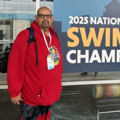 Nicholas Delgado poses next to a banner for the swimming competition at the 2025 National Senior Games. He's wearing a red hoodie, a backpack, and nametag.