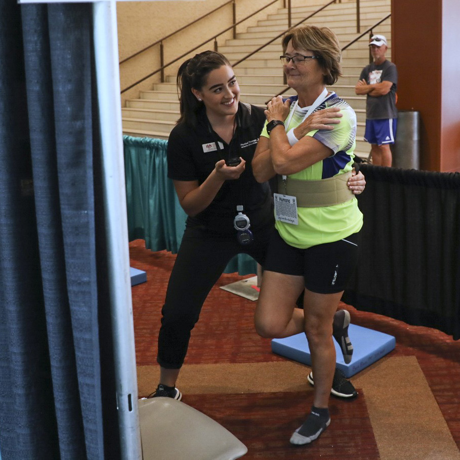 A National Senior Games athlete stands on one leg with their eyes closed and arms crossed across their chest. A physical therapy professional stands next to the athlete holding a stopwatch.