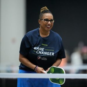A National Senior Games athlete smiles while standing ready to play on a pickleball court. The athlete is wearing a Humana Game Changer t-shirt. 
